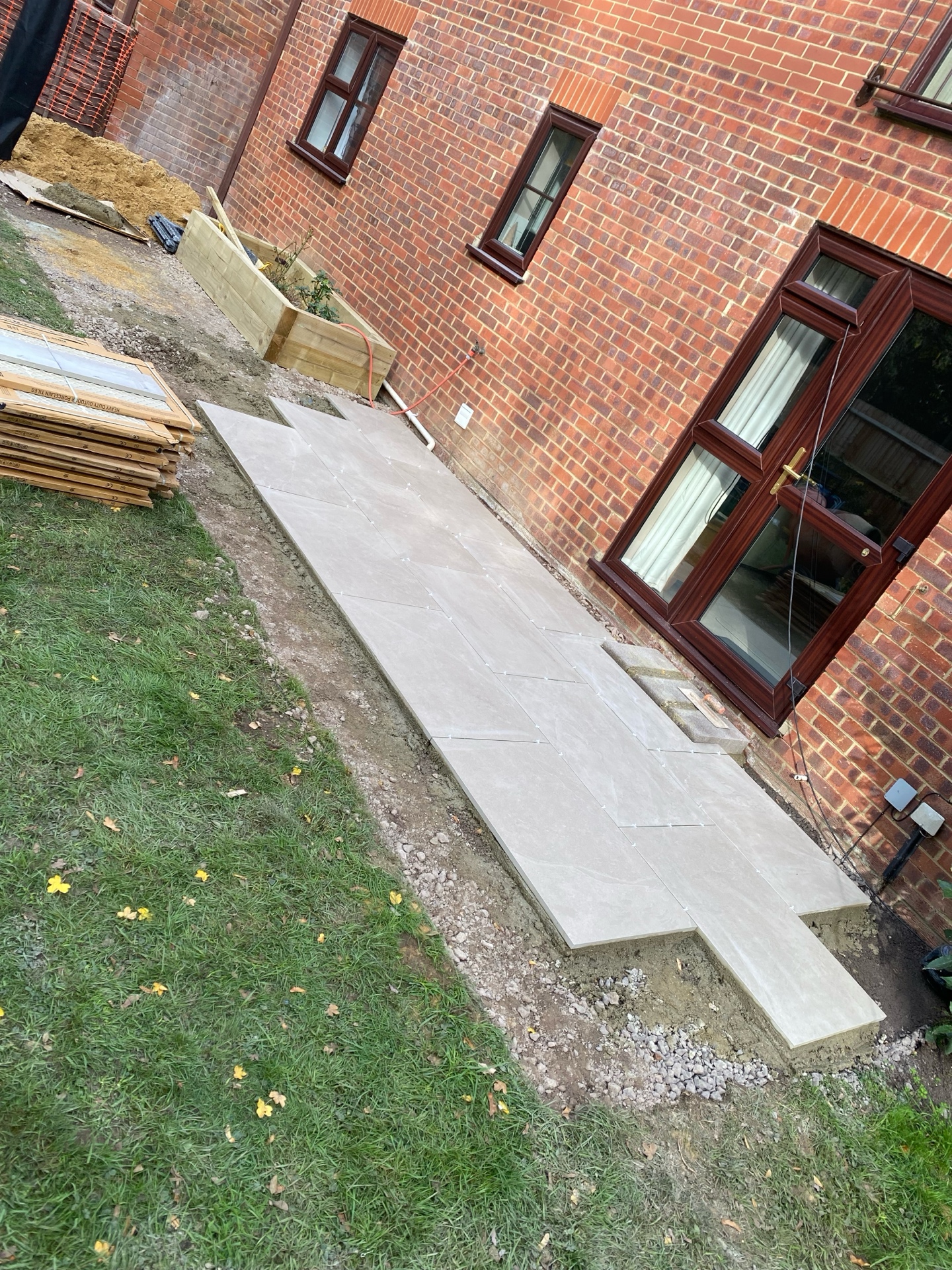 Patio paving in progress beside a brick house, with light-coloured paving slabs, prepared ground and a timber planter visible.