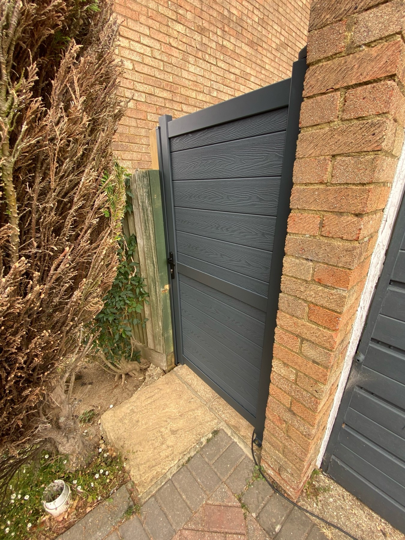 Completed anthracite-grey composite side gate with horizontal panels, dark aluminium frame and secure hardware beside brickwork.